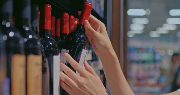 Close-up, a woman's hand takes a bottle of wine from the shelf. Purchase of an alcoholic beverage in the department of alcoholic beverages of the supermarket. Bottles stand in a row on the shelf of