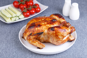 Chicken baked whole to a delicious crust. A traditional dish. In the background are cucumbers and cherry tomatoes on a white plate. Close-up, selective focus, gray background.