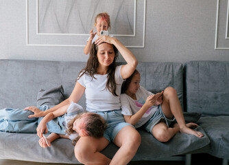 Tired young mother sits on couch and holds her head while her three naughty daughters play games on...