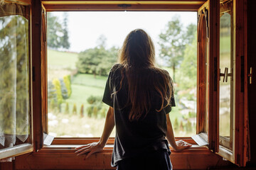Rearview of long dark-haired woman in t shirt looking out the window, contemplating landscape rustic natural tree view - Powered by Adobe