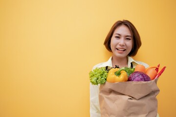 Happy vegetarian young Asian woman 20s in casual clothes hold paper bag with vegetables on workspace area copy space mock up isolated on Yellow background Shopping concept