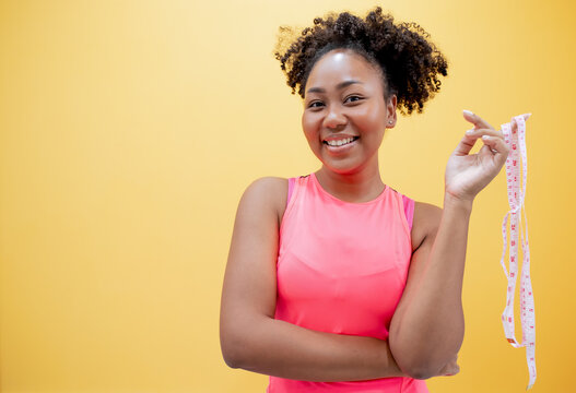 Smiling beautiful confused young African plus size body positive woman 20s wearing Sport wear hold measure tape looking camera isolated on yellow color wall background studio portrait.