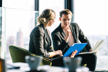 Two business man and woman talkin to each other discussing their business proposal. Both wearing white shirt and black jacket. They are sitting on the wooden table full of glass.