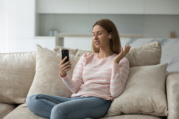 Positive pretty adult millennial mobile phone user woman talking on video call, using small wireless earphone, holding smartphone, resting on sofa at home, enjoying leisure, communication