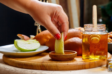 A woman's hand dips a piece of apple in honey in honor of the celebration of Rosh Hashanah near honey and challah and traditional food.