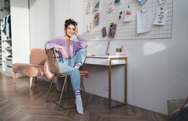 Young cute smiling woman sitting near table with laptop computer in home interior. Carefree female student enjoying distantly learning with modern technology and modern device