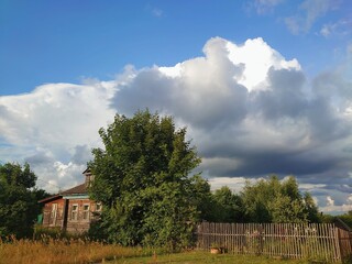 Country summer landscape with wooden house, blue sky and fluffy white clouds.