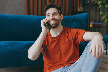 Young fun man wears red t-shirt talk speak on mobile cell phone sit on blue sofa couch stay at home...