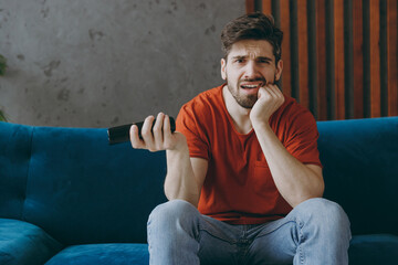 Young sad man wears red t-shirt hold TV remote controller watch film switch channels sit on blue sofa couch stay at home hotel flat rest relax spend free spare time in living room indoors grey wall.