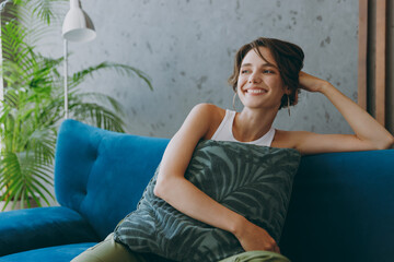 Young smiling woman wear white tank shirt hold pillow look aside sit on blue sofa couch stay at home hotel flat rest relax spend free spare time in living room indoors grey wall People lounge concept