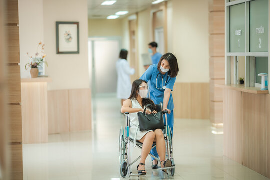 Smiling young female doctor in scrub and covid-19 face mask in conversation with patient sitting in wheelchair holding purse during discharge from hospital