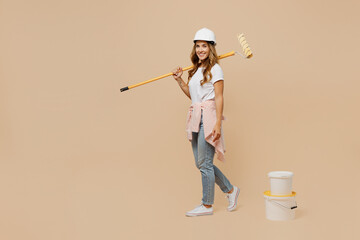 Full body young happy employee handyman woman in white t-shirt helmet hold paint roller walk near bucket isolated on plain beige background Instruments accessories for renovation room. Repair concept.