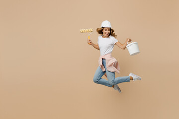 Full body young employee laborer handyman woman in white t-shirt helmet jump high hold paint roller...
