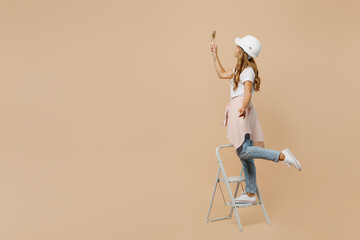 Full size young employee laborer handyman woman in white t-shirt helmet use ladder hold paint brush...