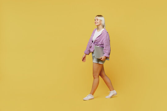 Full body side view young programmer student blonde IT woman 20s wear pink tied shirt white t-shirt hold closed laptop pc computer walk go isolated on plain yellow background People lifestyle concept - Powered by Adobe