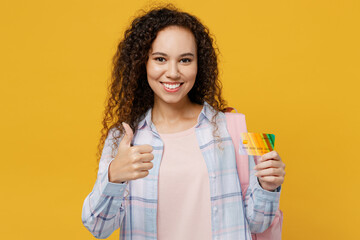 Young black teen girl student she wear casual clothes backpack bag hold in hand mock up of credit bank card show thumb up isolated on plain yellow background. High school university college concept.