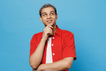 Young smiling fun minded man of African American ethnicity 20s he wear red shirt look aside on workspace area prop up chin isolated on plain pastel light blue cyan background People lifestyle concept