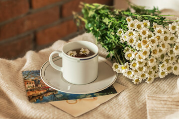 Morning coffee in the interior. Cozy composition: a cup of coffee with a saucer on a warm sweater and a bouquet of wild flowers against a brick wall. Still life concept. Copy space.