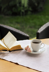 Morning coffee. Cozy interior: a cup of coffee and an open book on the table against the background of a wicker chair. Relax on the terrace in the garden.