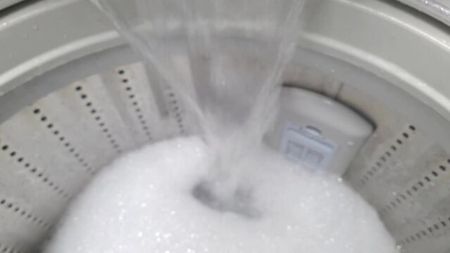 The top loading washing machine is working. Close-up white foam with bubbles on surface of water, washing powder.