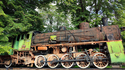 old abandoned and rusty passenger train in a spooky forest with the wheels on the rail, trees and greenery around - steampunk scene of vintage machinery outdoors