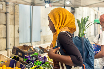 Young Black Muslim woman in hijab buying vegetables on a market street food, African Islamic lady with backpack outdoors 