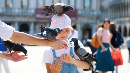 VENICE, ITALY - JULY 7, 2018: view of happy kid girl, tourist, holding pigeons, feeding, play with them, having fun on Piazza San Marco, St Mark's Basilica, on a summer day. High quality photo