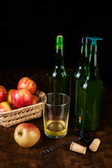 Bottles of cider, one with a stopper to pour next to a bottle opener, a glass with cider and some apples, on a dark background and a wooden table.
