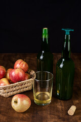 Bottles of cider, one with a stopper for pouring, next to a glass of cider and some apples, on a dark background and a wooden table.