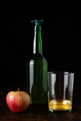 Bottle of cider with a stopper to pour next to a glass with cider and an apple, on a dark background and a wooden table.