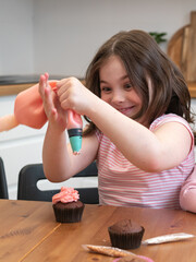 Funny little Brunette girl in the kitchen with disheveled hair looks at a cupcake, squeezes pink cream out of a pastry bag, grimaces. A master class on cooking for children