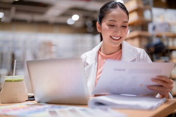 asian female smart entrepreneur working use laptop smartphone manage and checking product inventory in warehouse showroom,asia woman smiling cheerful business owner work in showroom warehouse store