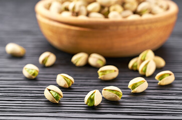 pistachios spill out of a wooden bowl on a black table.