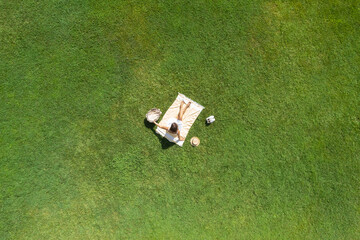 Woman in a white dress and hat, sitting on the plaid on the green grass having a picnic with food...