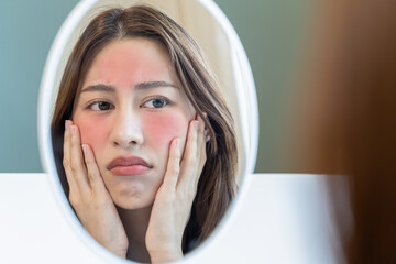 Asian young woman looking face skin in the mirror have a red rash on her face from cosmetic allergy