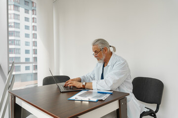 Fototapeta na wymiar Focused male healthcare worker doing paperwork and using laptop while working at doctor's office