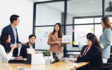 Asian beautiful business woman wearing formal suit, discussing and presenting financial graph or chart to her mixed race colleagues in meeting room at indoor office or workplace.