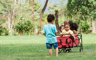 Obraz premium Mixed race little cute children pull and drag a red cart while playing in outdoor green park. Education, Diversity and Teamwork Concept.