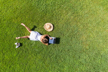 Woman in a white dress, hat with laptop, lying on the green grass at summer day. Top view, drone,...