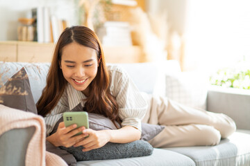 Smiling asian young woman relax on sofa and texting messaging on smartphone,browse wireless internet on mobilephone.Happy girl use cellphone chatting with her friend,video call,sending email at house