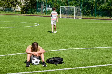 Sad disappointed teenage boy sitting in empty school sport stadium outdoors. Friend walks away and leaves his friend sitting alone. Emotions, defeat, lost game, difficulties, problems of teenagers