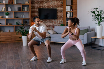 Cheerful young african american husband and wife in sportswear doing squat exercises
