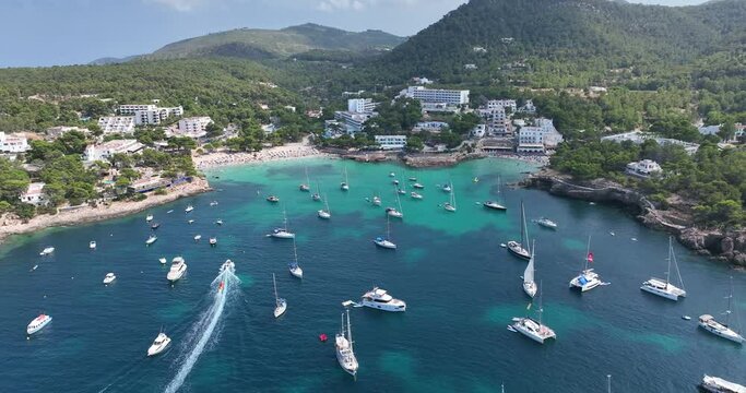Paradise beach surrounded by trees. Yachts and boats anchored in front of the shore where people enjoy the sun.