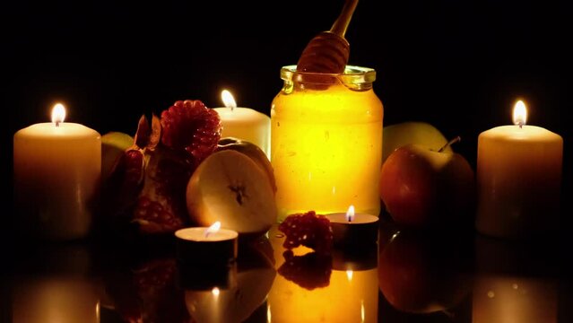 Golden honey, fresh apples and pomegranate on black background. Burning candles. Happy Rosh Hashanah concept. Wooden spoon for honey in human hand. Traditional food of jewish new year celebration.