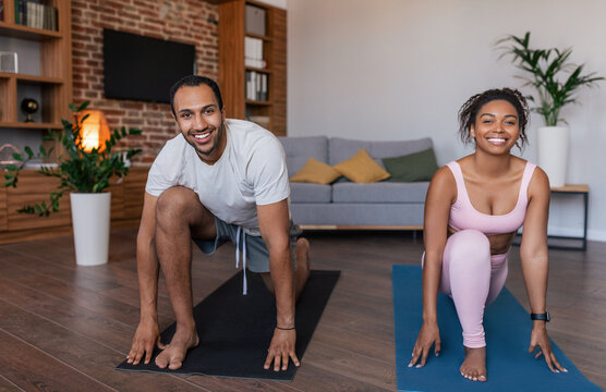 Smiling young black man and lady in sportswear doing leg exercises, stretching or practicing yoga together