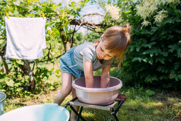 Naklejka na ściany i meble Little preschool girl helps with laundry. Child washes clothes in garden