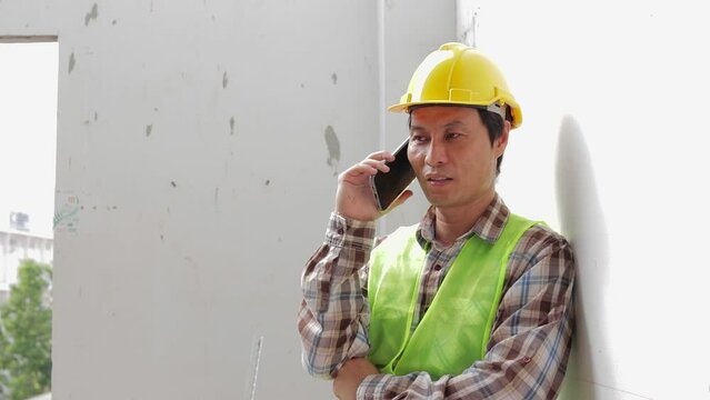 A construction worker wearing a yellow helmet holds a smartphone talking to the coordinator at the construction site. Construction industry concept. real estate project