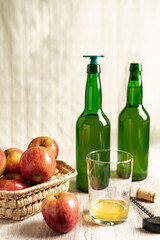 Glass with a little cider, next to some bottles and some apples, on a light wood table.