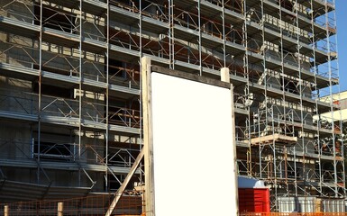 Blank information board outside the construction site with a new concrete building among scaffolding