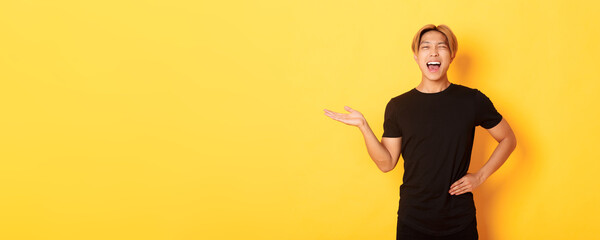 Portrait of happy blond asian guy, winking sassy and smiling, holding something on hand over yellow...